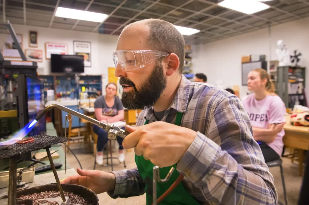 A bearded man wearing safety goggles and a plaid shirt uses a blowtorch on a workbench in a workshop, while three people sit in the background watching attentively.