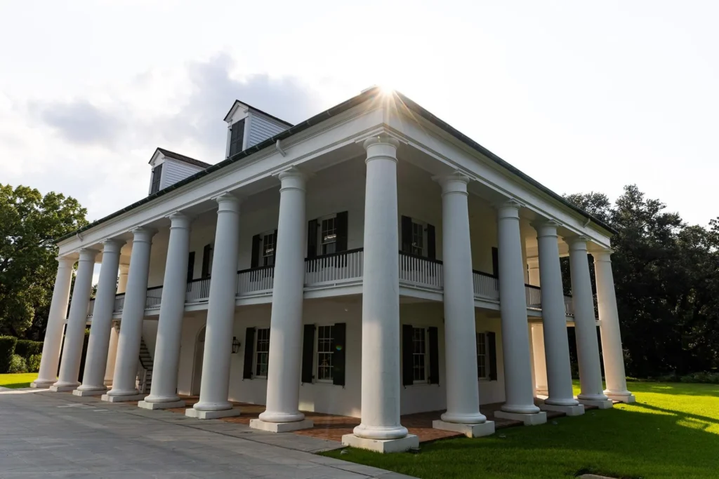 Large white plantation-style house with tall columns, a wide front porch, and two stories; sunlight shines through the roof, and green lawn surrounds the building. Trees are visible in the background.