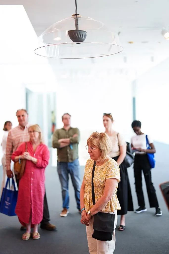 A group of people stands in an art gallery, observing an exhibition. A woman in a yellow top is in the foreground, and a large, clear dome-shaped speaker hangs from the ceiling above her.
