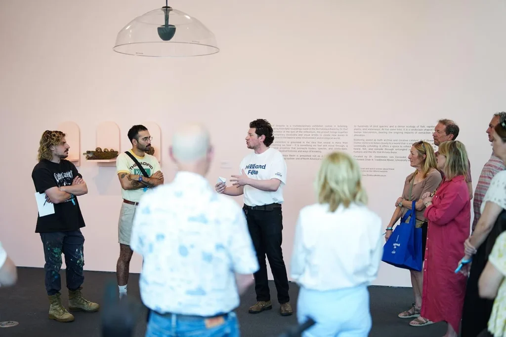 A group of people listens to a man speaking in front of a white wall with text and small artworks, in a brightly lit gallery space. Some attendees hold bags and notebooks, while others stand with arms crossed.