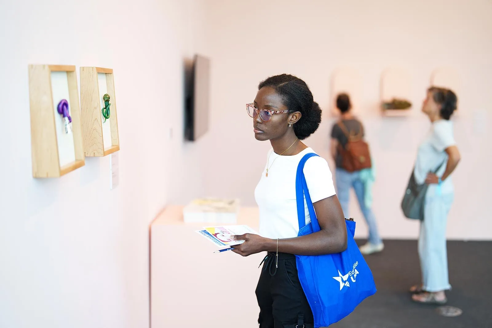A woman with glasses and a blue tote bag looks at framed artworks on a gallery wall, holding papers. Two other visitors view displays in the background. The room is bright and minimalistic.