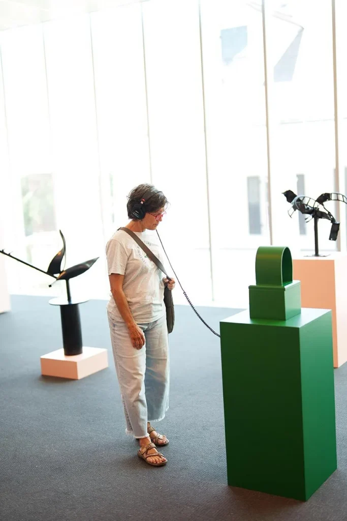 A woman wearing headphones stands in a bright gallery, listening to an audio guide while observing a green abstract sculpture on a pedestal. Other sculptures are displayed in the background.