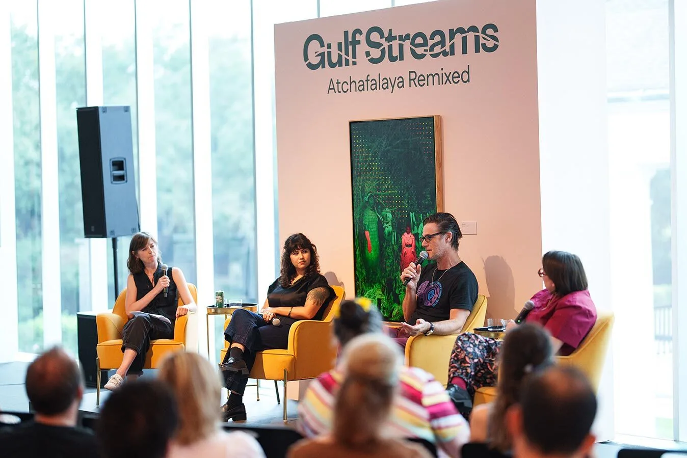 Four people, including Rachel Libeskind, sit on yellow chairs before an audience, speaking in a panel discussion. Behind them is a sign reading Gulf Streams: Atchafalaya Remixed and a painting of green plants and a pink bird.