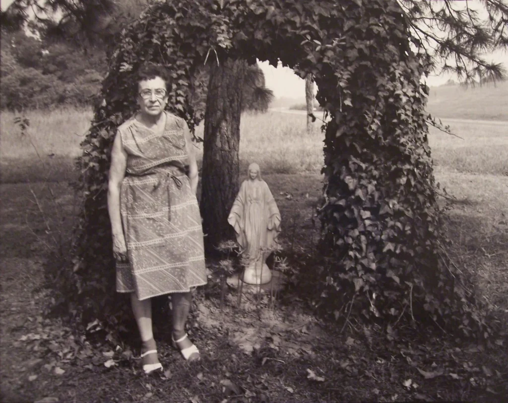 An older woman in a patterned dress stands beside a tree covered in ivy, with a small statue of Jesus beneath the arch of vines, in a grassy outdoor setting.