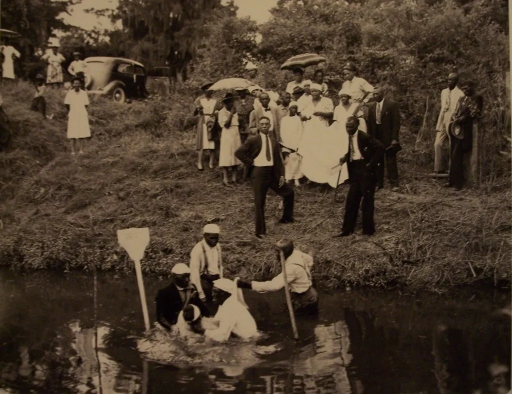 A black-and-white photo of a group of people in formal clothes gathered by a riverbank. Several individuals are in the water, performing a baptism while others watch from the grassy shore, some holding umbrellas.