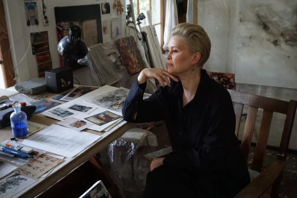 A woman with short blonde hair, dressed in black, sits thoughtfully at a cluttered art studio desk covered with papers, art supplies, and photographs. Artworks and materials are visible in the background.