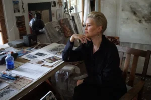 A woman with short blonde hair, dressed in black, sits thoughtfully at a cluttered art studio desk covered with papers, art supplies, and photographs. Artworks and materials are visible in the background.