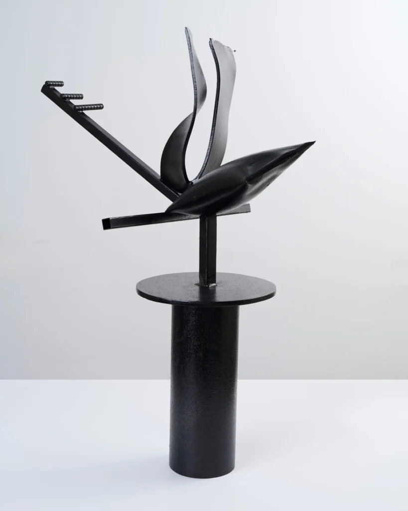 A modern black metal abstract sculpture stands on a cylindrical pedestal, featuring curved and angular elements branching out in different directions, set against a plain light background.