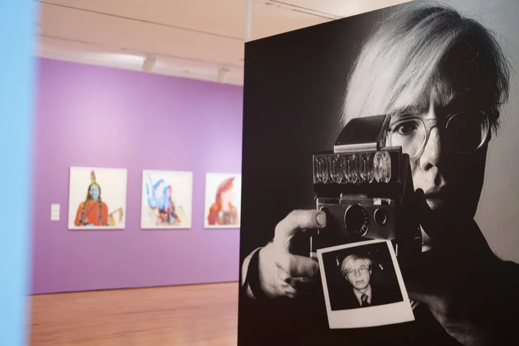 A large black-and-white photo of a man holding a camera and a Polaroid is displayed in a gallery with purple walls and colorful framed artworks in the background, capturing the spirit of Resonant Roots & The Art of Listening.