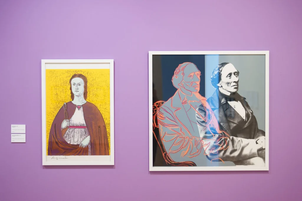 Two framed portraits hang on a purple wall: the left is a stylized illustration of a woman in a red cape, and the right is a composite image of a man, one side in grayscale, the other in neon outlines.