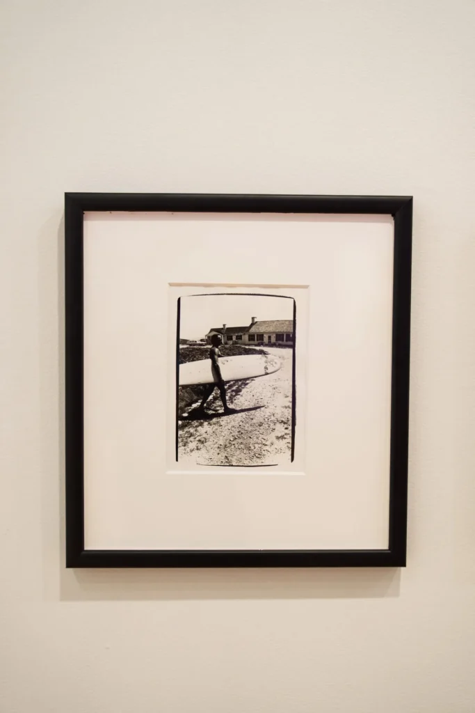 Black-and-white photo in a black frame shows a person walking on a curved path toward a house in the background, displayed on a plain, light-colored wall.