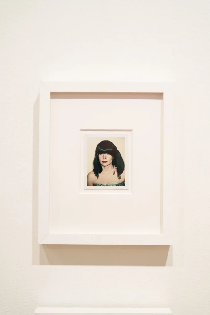 A framed photograph of a woman with dark hair and bangs, wearing a strapless top, displayed on a plain white wall.