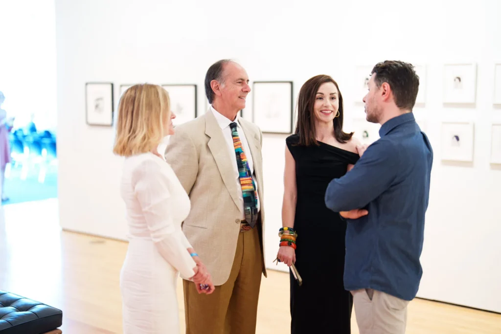 Four adults stand in a brightly lit art gallery, smiling and talking together. Framed artwork hangs on the white walls behind them. The group appears engaged and well-dressed, suggesting a formal event or exhibition.