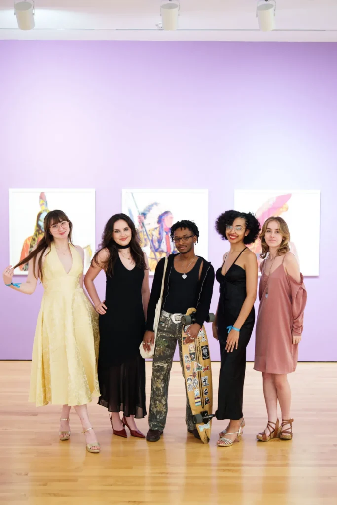 Five people in stylish dresses and heels pose together in an art gallery with a purple wall and colorful artworks behind them. One person holds a skateboard. The group is smiling and standing on a wooden floor.