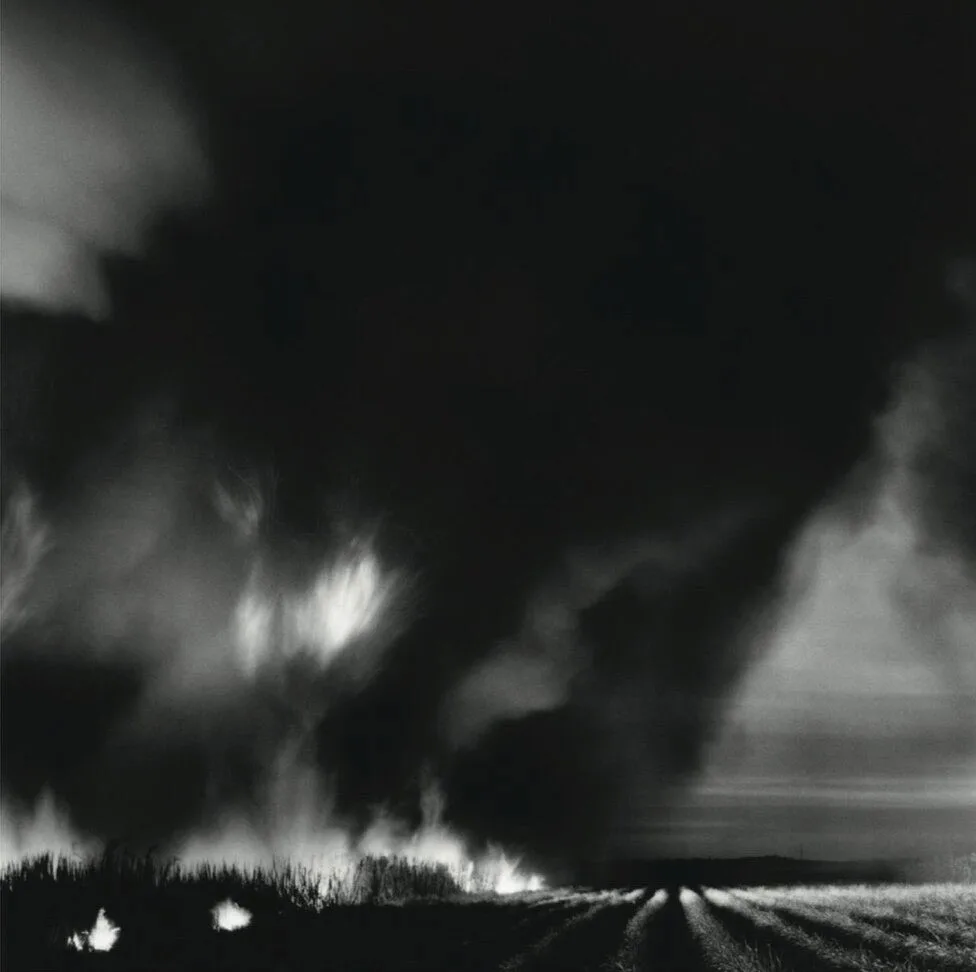 Thick, dark smoke billows upward from a fire burning fields, with flames at the base and crop rows visible in the foreground under a dramatic sky—a scene reminiscent of the evocative landscapes often explored by Matthew Day Jackson.