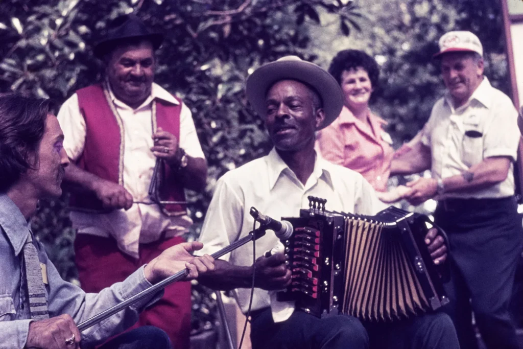 A group of people play music outdoors; one person plays accordion, another plays a washboard, while others smile and watch. Trees are visible in the background.