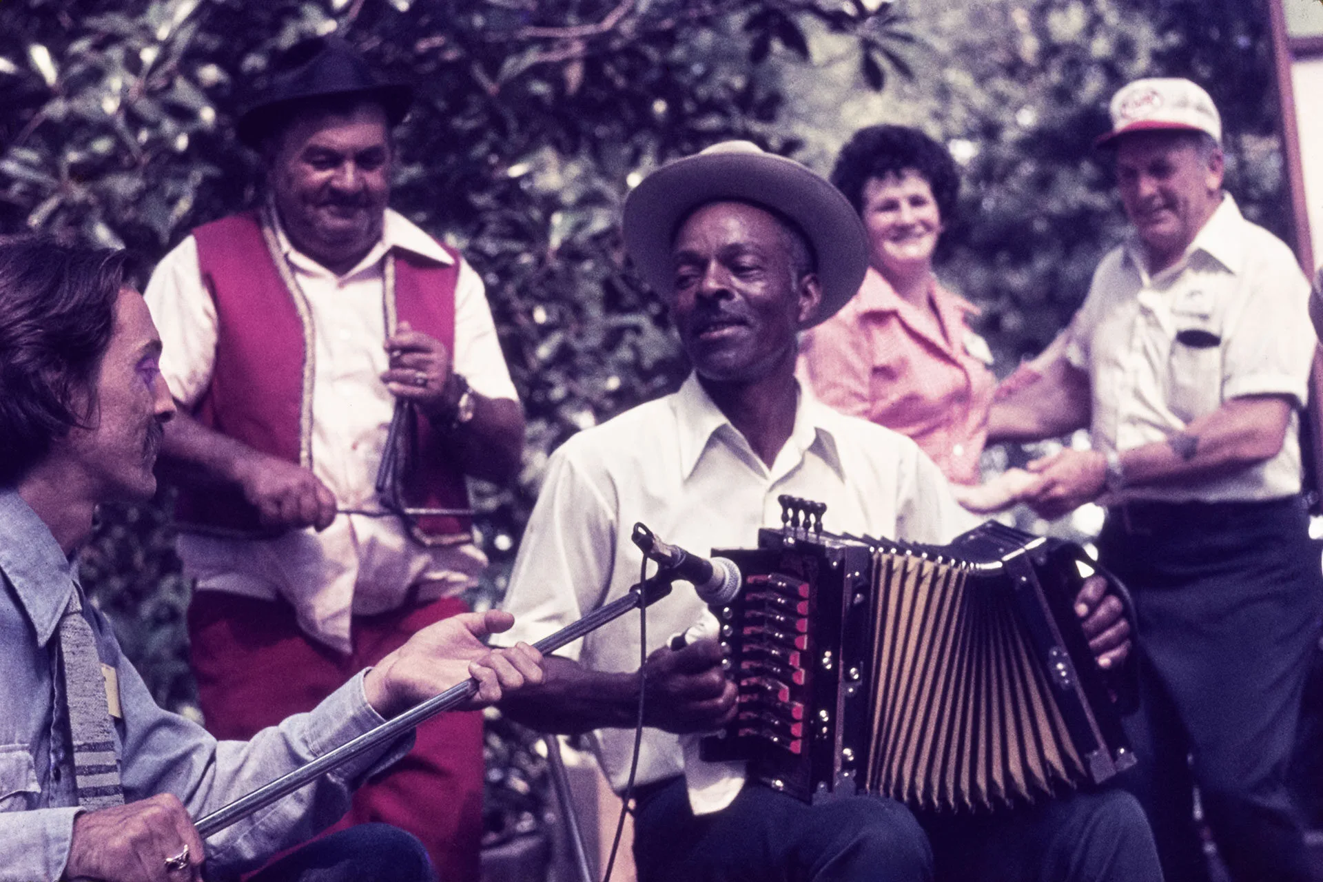 A group of people play music outdoors; one person plays accordion, another plays a washboard, while others smile and watch. Trees are visible in the background.