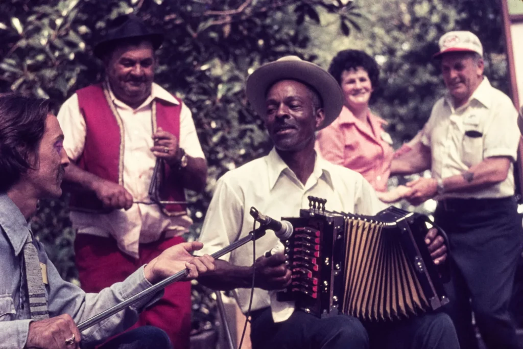 Five people play music outdoors; one plays accordion, another fiddle, while others smile and watch. The group includes men and a woman, all casually dressed. Trees and greenery are visible in the background.