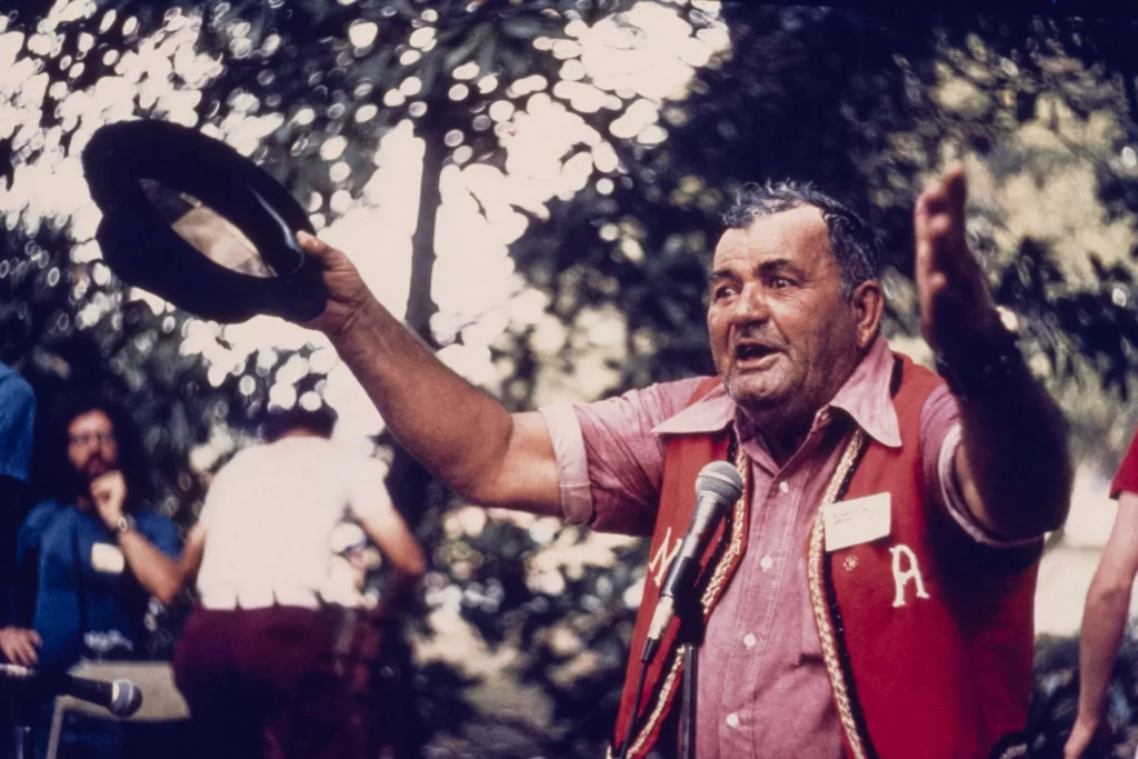 An older man in a red vest holds a hat in one hand and gestures with the other while speaking into a microphone outdoors. Trees and people are visible in the background.