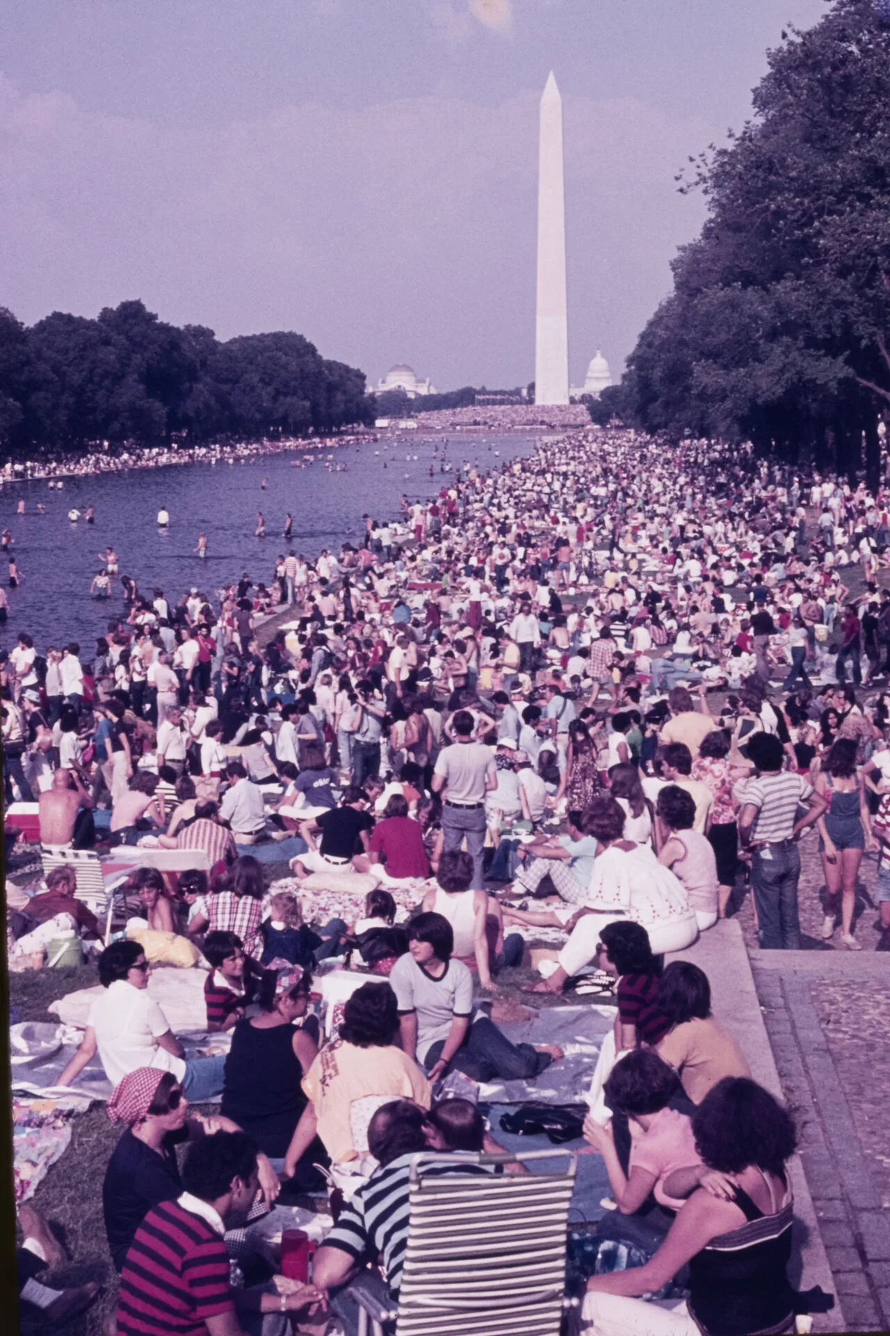 Large crowd gathers by the Reflecting Pool in Washington, D.C., with the Washington Monument in the background on a clear day. Many people are sitting, standing, or wading in the water.