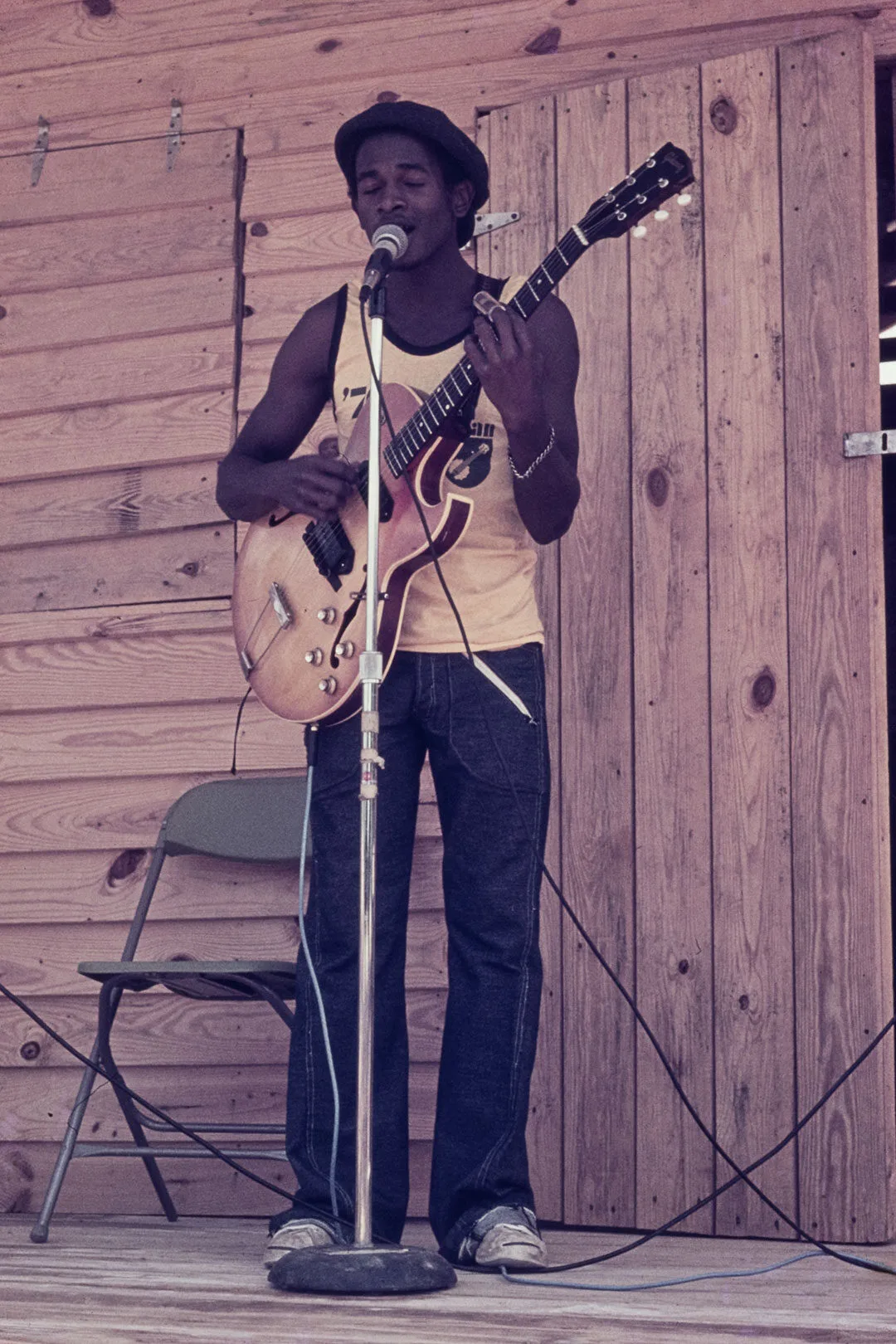 A man wearing a hat and tank top plays an electric guitar and sings into a microphone on an outdoor wooden stage beside a folded chair.