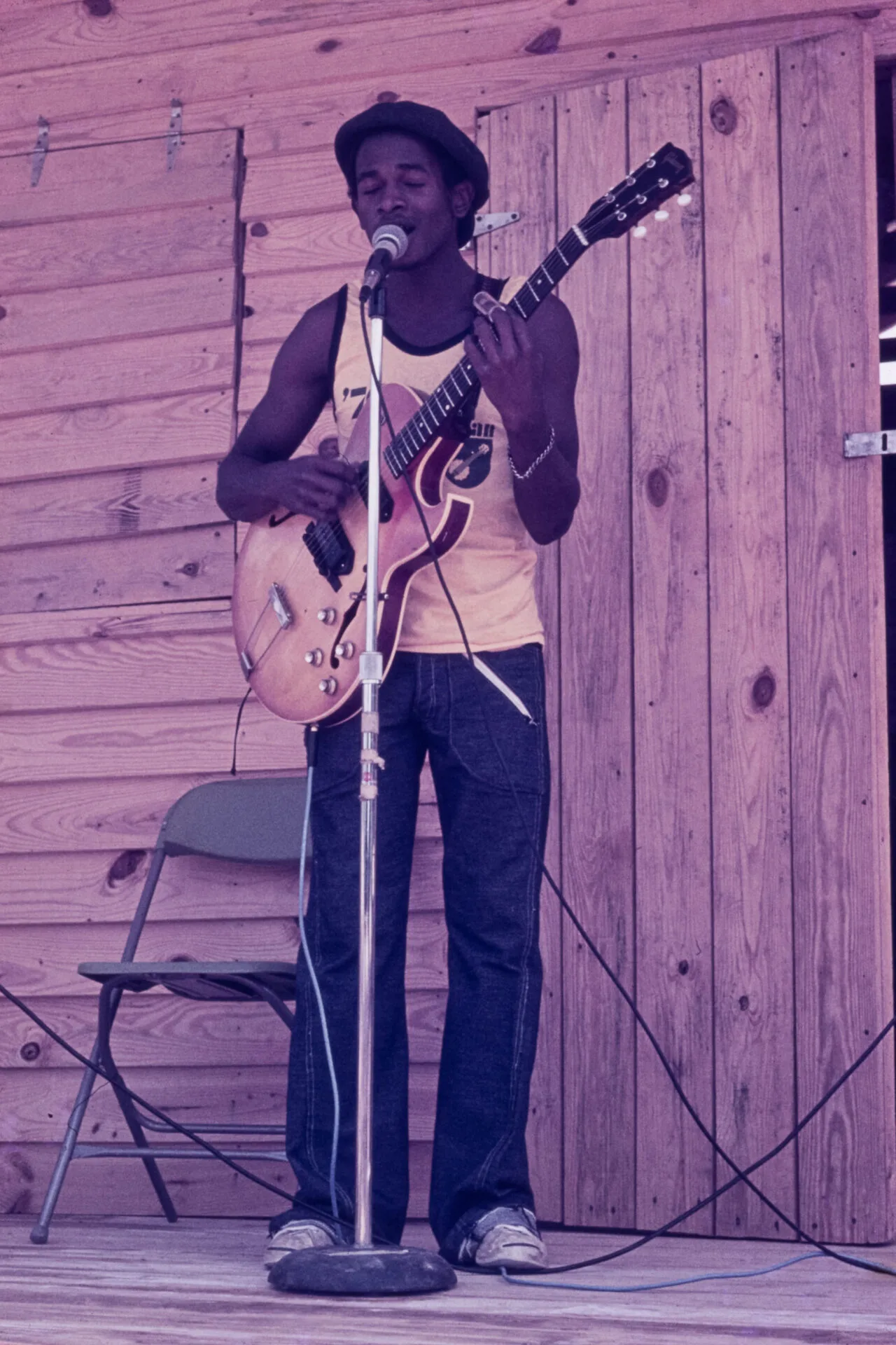 A musician wearing a hat and tank top plays an electric guitar and sings into a microphone on a wooden stage, with a chair and wooden wall in the background.