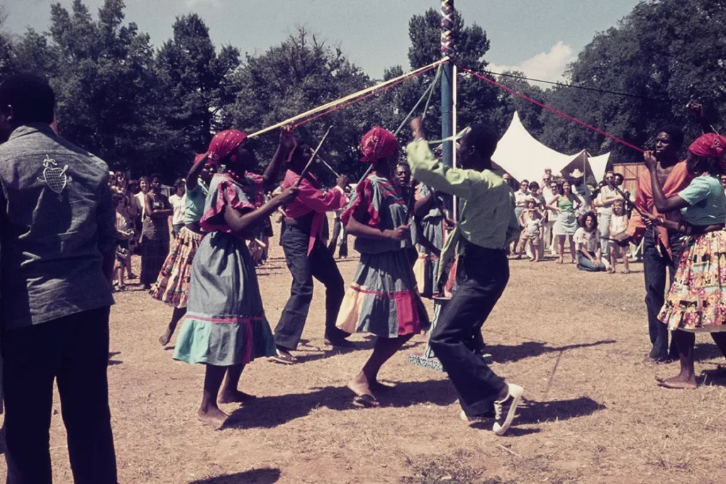 People in colorful outfits dance around a maypole, weaving ribbons, while a crowd watches outdoors. Trees and a white tent are visible in the background on a sunny day.