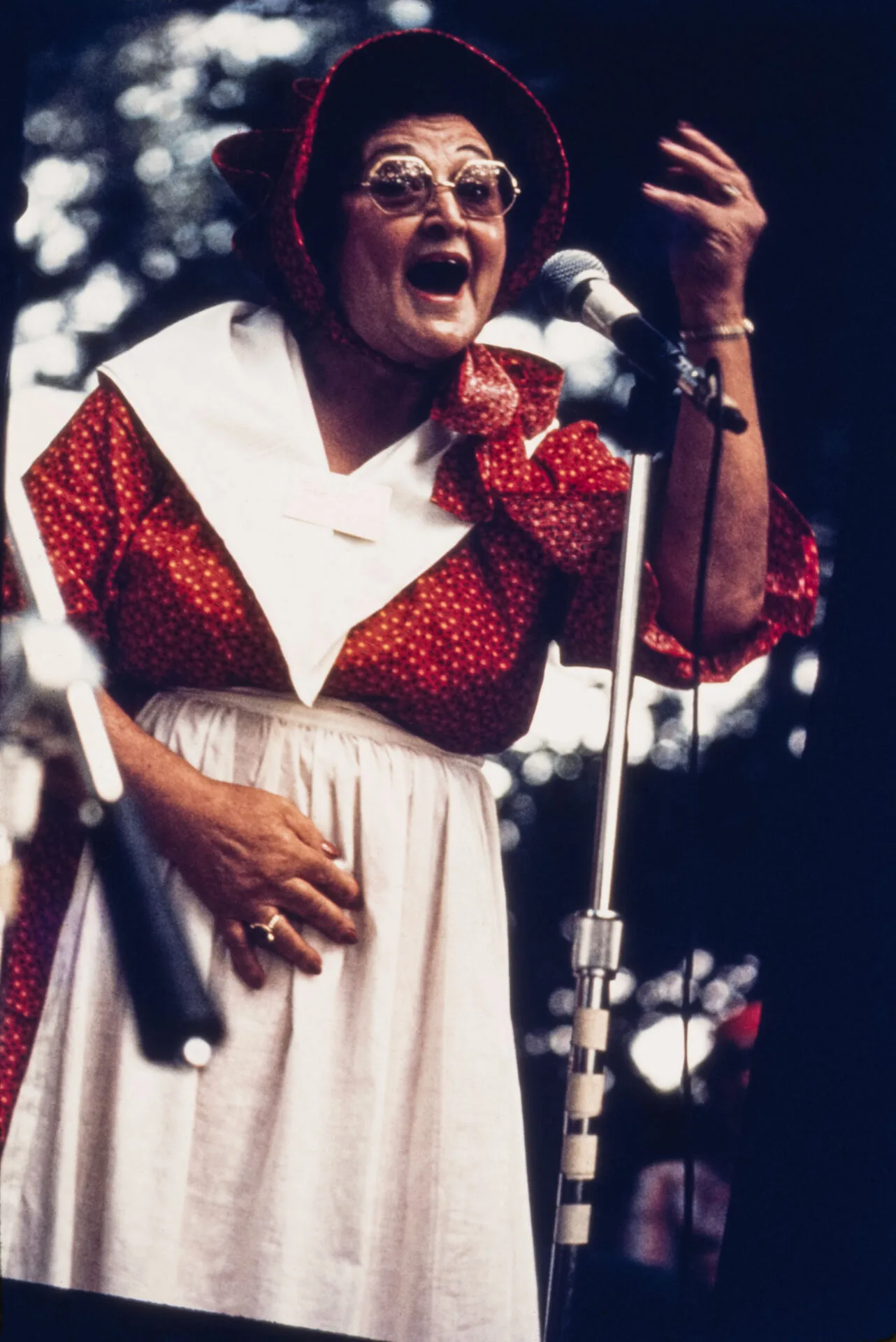 A person wearing a red bonnet, red dress with white polka dots, and a white apron passionately sings or speaks into a microphone on stage, evoking the vibrant spirit often captured in the works of Elemore Morgan Jr.