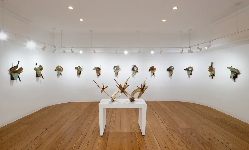 A modern art gallery with wooden floors displays abstract metal sculptures on white walls and a white pedestal, all under bright ceiling lights, reminiscent of the striking installations inspired by Elemore Morgan Jr.
