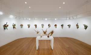 A modern art gallery with wooden floors displays abstract metal sculptures on white walls and a white pedestal, all under bright ceiling lights, reminiscent of the striking installations inspired by Elemore Morgan Jr.