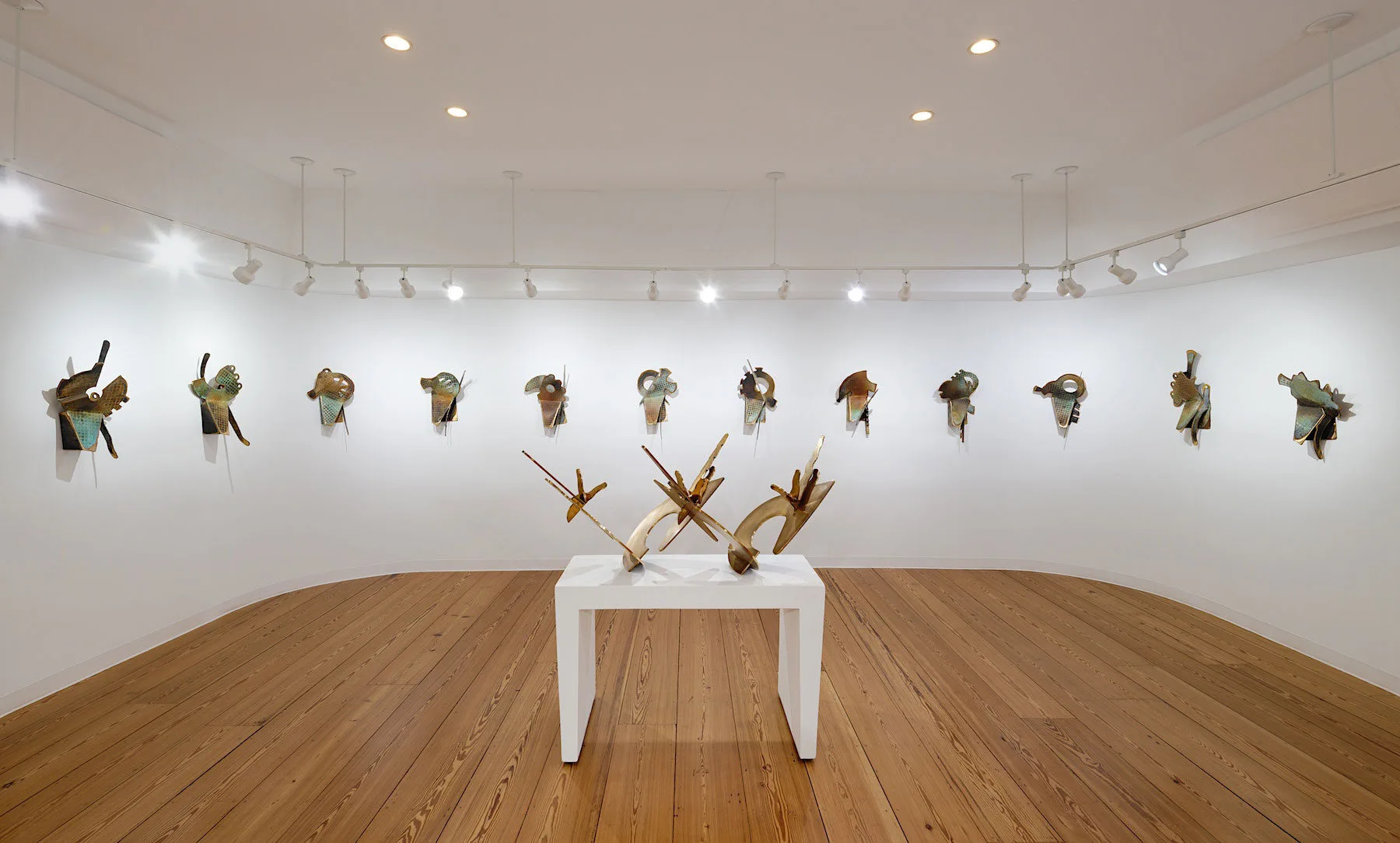 A modern art gallery with wooden floors displays abstract metal sculptures on white walls and a white pedestal, all under bright ceiling lights, reminiscent of the striking installations inspired by Elemore Morgan Jr.
