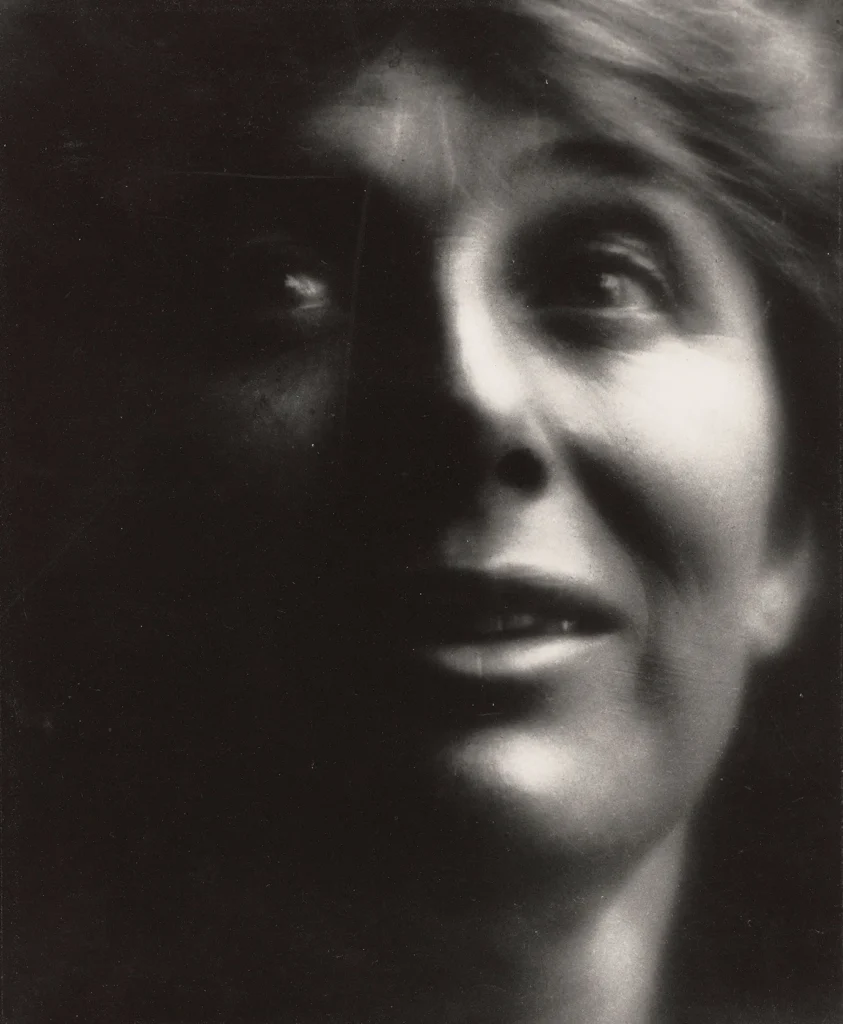 A black and white photo of a person’s face, half-lit and half in shadow, with a slight smile and eyes looking upward. Reminiscent of Matthew Day Jackson’s evocative portraiture, the image has a soft focus and dramatic lighting.