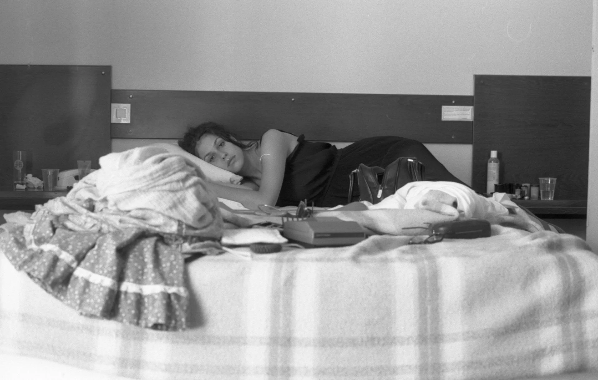 A woman lies on her side in bed, resting her head on a pillow and looking toward the camera. The bed is unmade and cluttered with clothes and items, evoking a scene reminiscent of Matthew Day Jackson’s explorations of everyday environments. The room is dimly lit and has a simple, minimal decor.