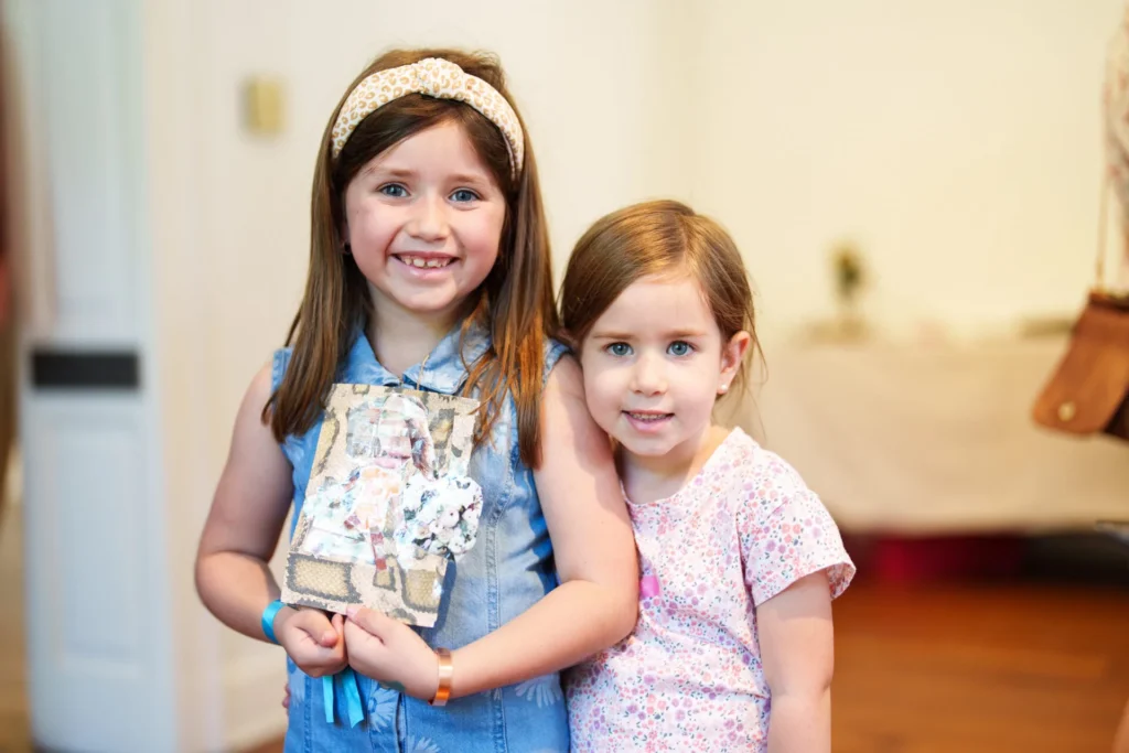 Two young girls stand indoors, smiling at the camera. One wears a headband and denim dress, holding a decorated journal. The other girl stands close beside her, wearing a pink patterned shirt. The background is softly blurred.