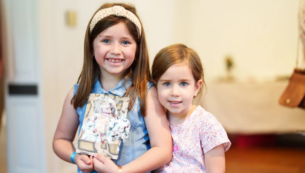 Two young girls stand indoors, smiling at the camera. One wears a headband and denim dress, holding a decorated journal. The other girl stands close beside her, wearing a pink patterned shirt. The background is softly blurred.