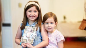 Two young girls stand indoors, smiling at the camera. One wears a headband and denim dress, holding a decorated journal. The other girl stands close beside her, wearing a pink patterned shirt. The background is softly blurred.
