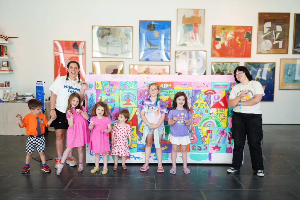 Seven children and two adults stand in front of a large, colorful art display. Artworks hang on the white wall behind them. The children smile, some holding hands, dressed in casual clothes.