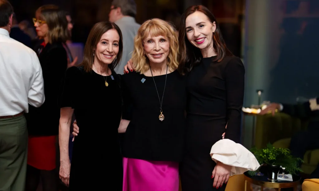 Three women dressed in black and pink pose together, smiling at an indoor event with other people socializing in the background.