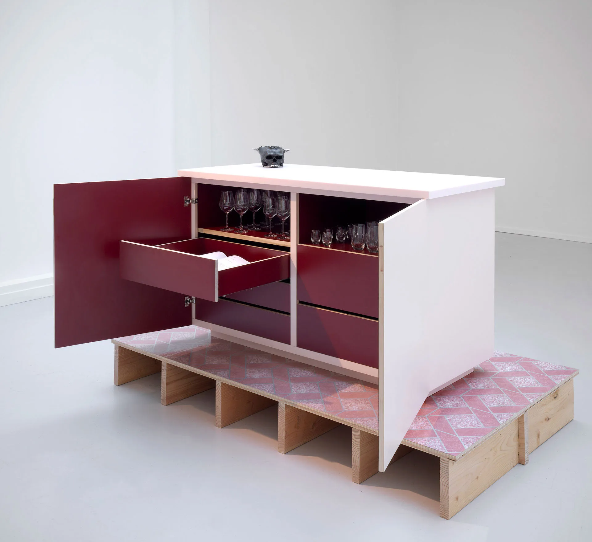 A white cabinet with maroon drawers and doors stands open, displaying shelves of glassware and books. It sits on a patterned platform made of plywood and pink tiles in a minimalist gallery space.