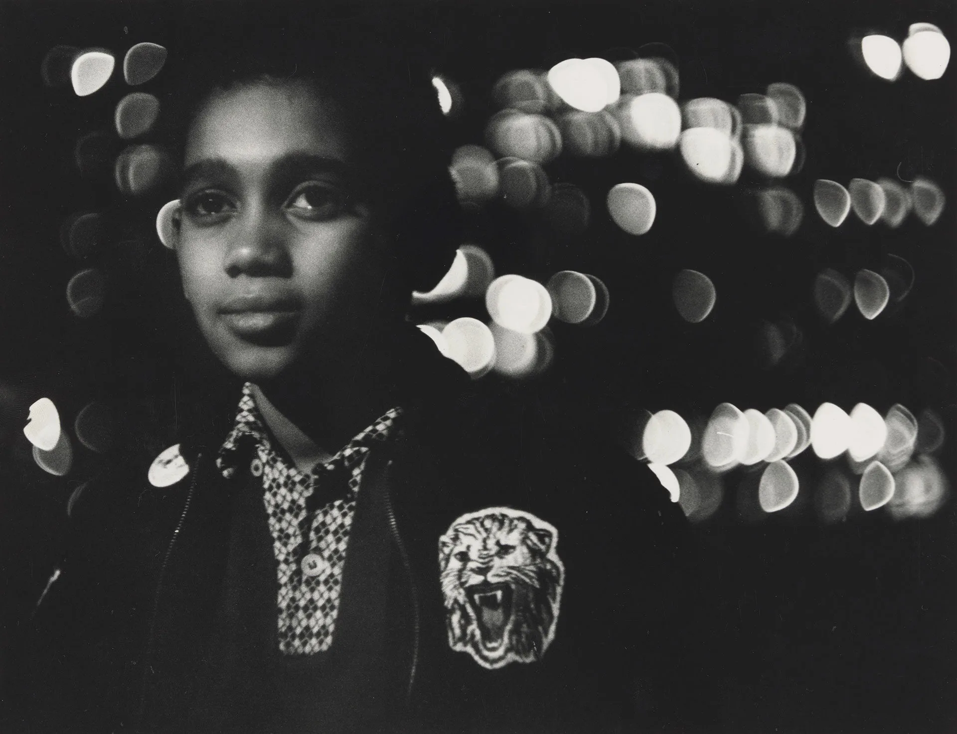A young person stands at night, wearing a jacket with a roaring tiger patch—a scene reminiscent of the evocative style found in Matthew Day Jackson’s artwork. The background is dark, with blurred, circular city lights creating a striking bokeh effect.