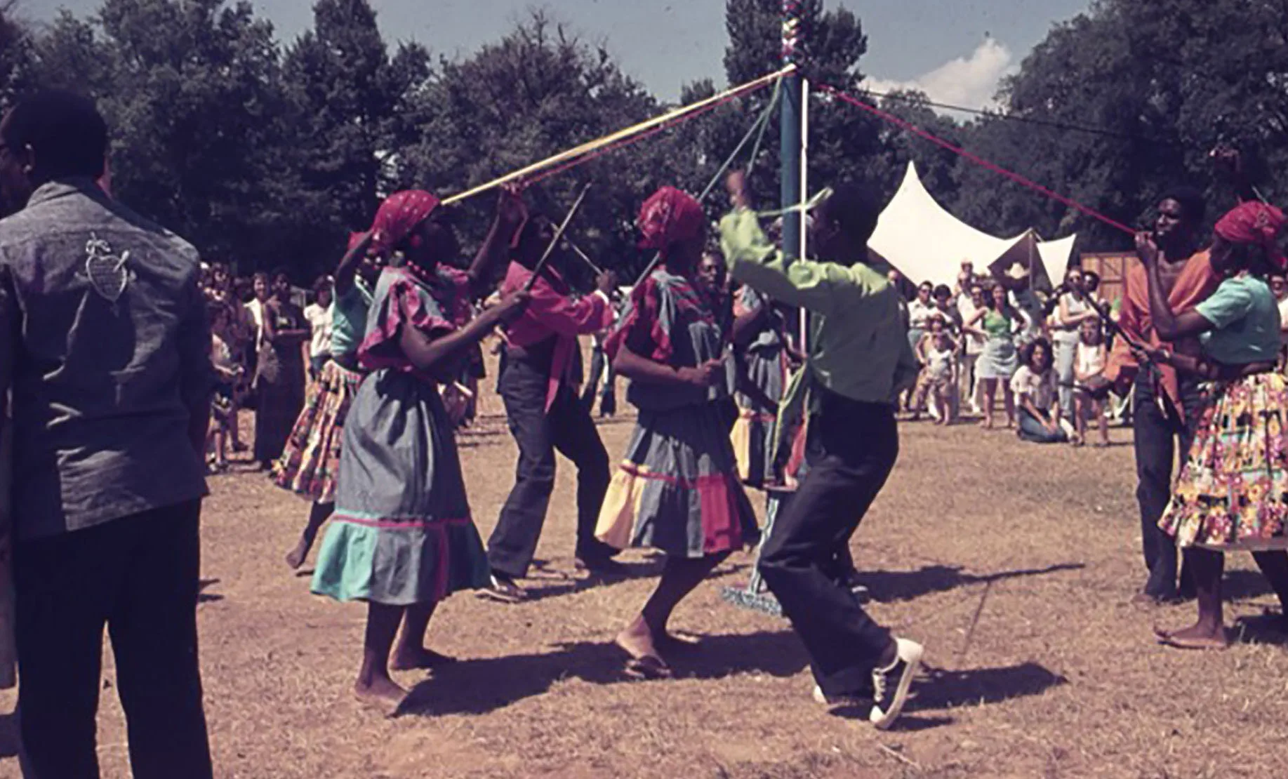 People in colorful clothing dance barefoot around a maypole, weaving ribbons, during an outdoor festival. Spectators watch in the background under trees and tents on a sunny day.