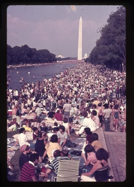Large crowd gathers around the Reflecting Pool in Washington, D.C., with the Washington Monument in the background, on a sunny day. People sit, stand, and relax, filling the area near the water.