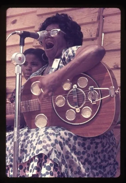 A woman in a patterned dress passionately sings and plays a resonator guitar at a microphone, with a child beside her. They are seated in front of a wooden wall.