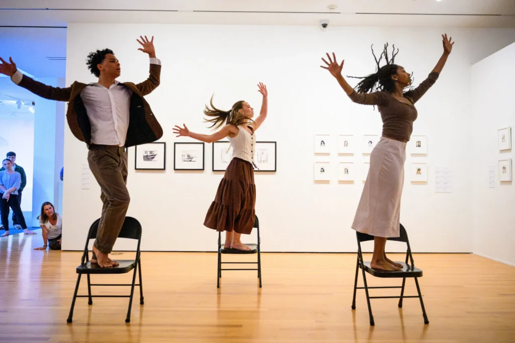 Three people stand on chairs with arms raised, dancing expressively in a museum gallery. Framed art is displayed on white walls behind them. A few onlookers watch from the corner.
