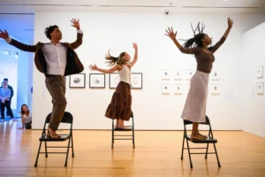 Three people stand on chairs with arms raised, dancing expressively in a museum gallery. Framed art is displayed on white walls behind them. A few onlookers watch from the corner.