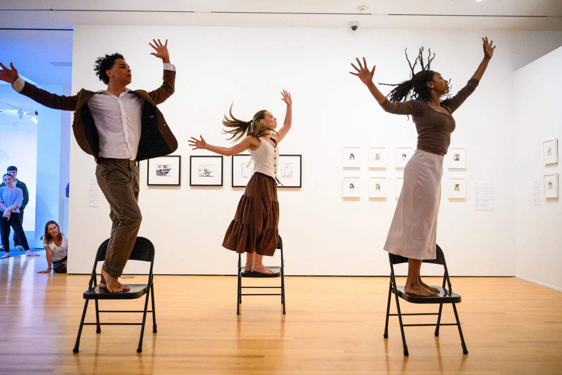 Three people stand on chairs with arms raised, dancing expressively in a museum gallery. Framed art is displayed on white walls behind them. A few onlookers watch from the corner.