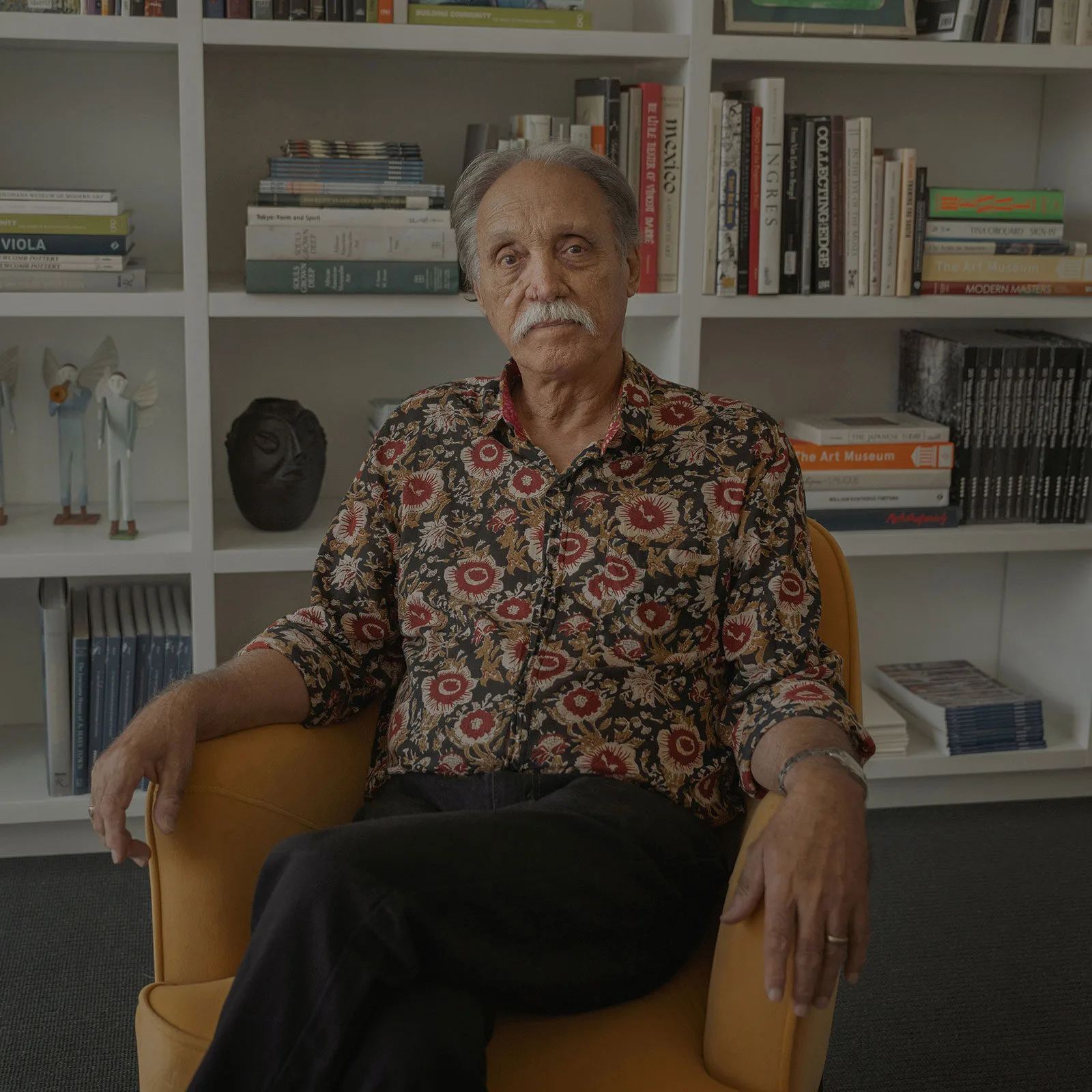 An older man with a mustache, wearing a floral print shirt, sits in a yellow armchair in front of a bookshelf.