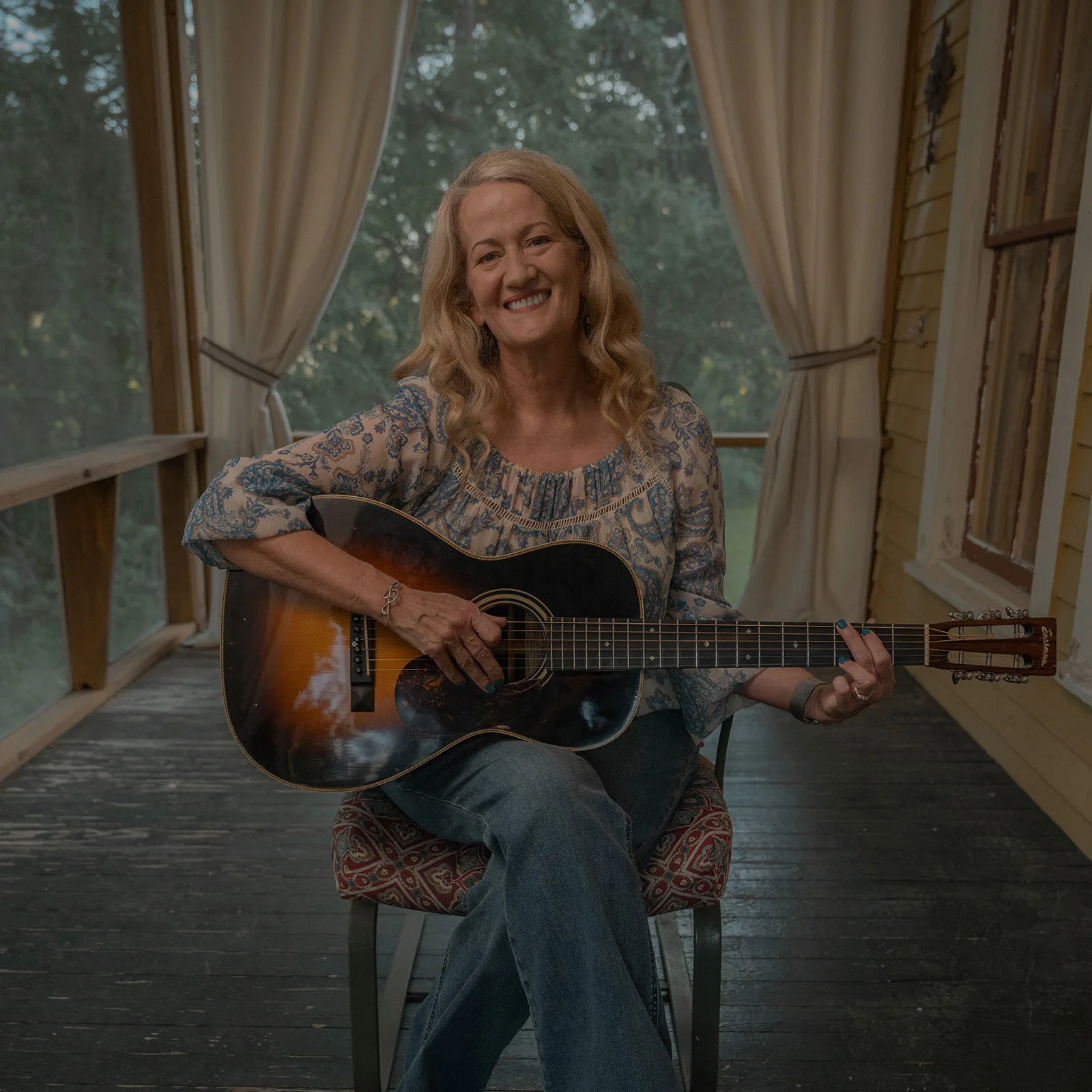 A smiling woman sitting on a porch playing an acoustic guitar.