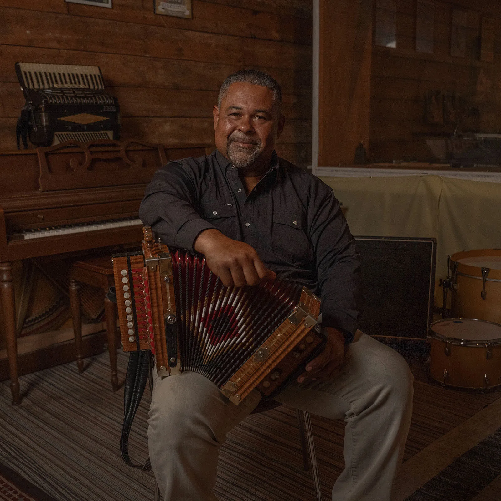 A man with a graying beard sits holding an accordion in a room with a piano and drums.