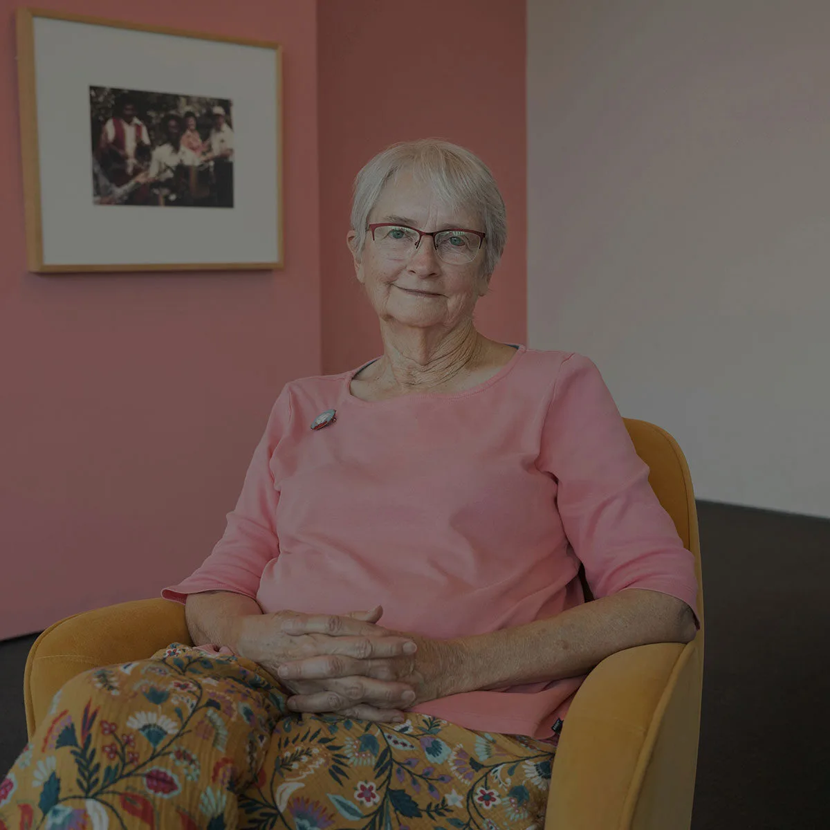 An older woman with short gray hair and red glasses sits in a yellow armchair in front of a pink wall with a framed photo.